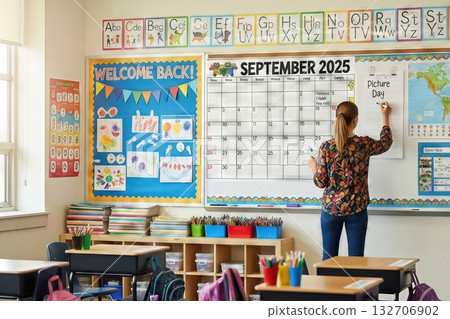 A teacher is updating a large classroom wall calendar for September 2025. The classroom is decorated with educational posters, supplies, and colorful displays under bright indoor lighting. The 132706902