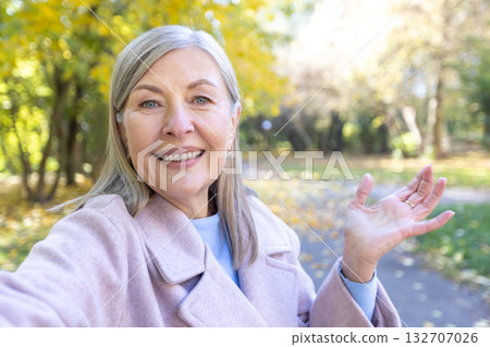 Senior woman smiling and waving hand during a video call or making a selfie video in an autumn park, embracing digital connectivity and communication with friends or family 132707026