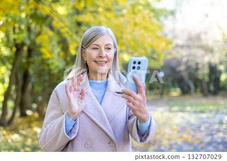 Senior woman smiling and waving during a smartphone video call in an autumn park, connecting with family or friends amid golden leaves and casual outdoor comfort Senior woman smiling and waving during a smartphone video call in an autumn park, connecting with family or friends amid golden leaves and casual outdoor comfort 132707029