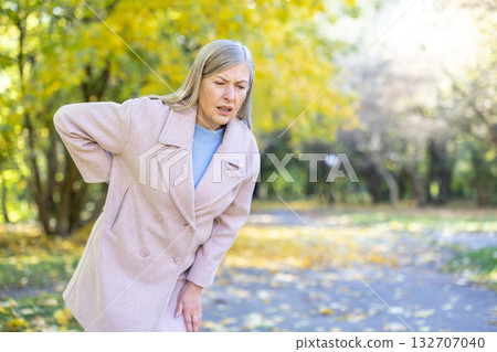 Senior woman standing bent over in an autumn park, suffering from acute lower back pain, clutching her side while showing a pained facial expression due to discomfort and health issues 132707040
