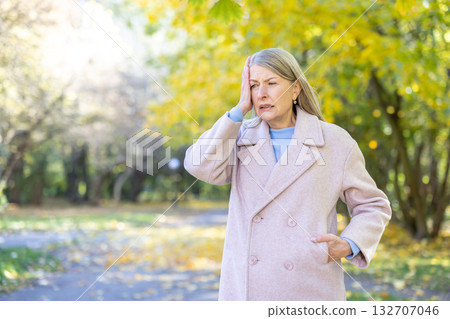 Senior woman suffering from a headache, holding her temple in a gesture of discomfort, indicating stress or unwellness while walking in an autumn park with colorful trees 132707046