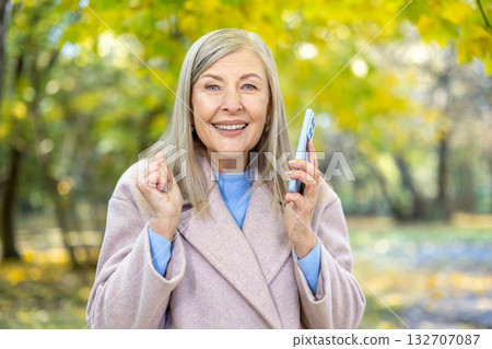 Senior woman is happily receiving good news during a phone call, expressing success and excitement with a clenched fist gesture in an autumn park setting 132707087