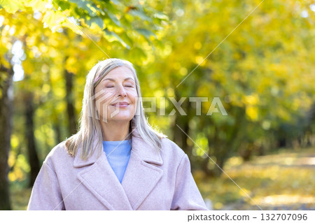 Senior woman relaxing with eyes closed and a gentle smile, feeling the sun on her face in an autumn park with blurred yellow foliage in the background Senior woman relaxing with eyes closed and a gentle smile, feeling the sun on her face in an autumn park with blurred yellow foliage in the background 132707096