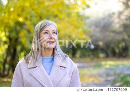 Mature woman with gray hair smiling and looking away, enjoying a peaceful autumn walk in a park surrounded by golden trees and soft natural light, feeling content and serene Mature woman with gray hair smiling and looking away, enjoying a peaceful autumn walk in a park surrounded by golden trees and soft natural light, feeling content and serene 132707099