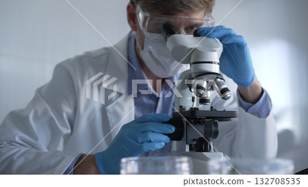 Male scientist researcher wearing blue gloves, protective glasses and medical mask, is using microscope in modern laboratory, portrait view. Low key lighting photo. Science and Medicine Male scientist researcher wearing blue gloves, protective glasses and medical mask, is using microscope in modern laboratory, portrait view. Low key lighting photo. Science and Medicine 132708535