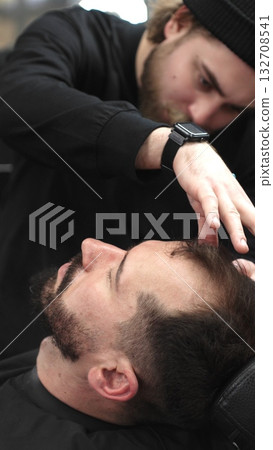 Professional barber carefully shaving client's beard with straight razor, applying gel and using hot towel for precise grooming in contemporary barbershop setting, vertical view 132708541