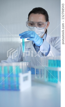 Female lab worker or scientist, wearing coat, blue medical gloves, mask and protective glasses is examining test tube with blue liquid in modern laboratory. Low key lighting photo Female lab worker or scientist, wearing coat, blue medical gloves, mask and protective glasses is examining test tube with blue liquid in modern laboratory. Low key lighting photo 132708543