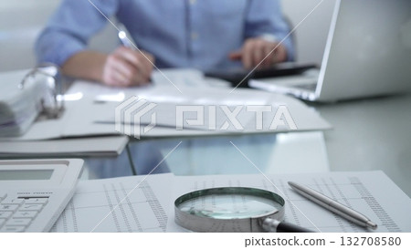 Magnifying glass, calculator, pen and documents are lying on accountant's desk. Businessman wearing blue shirt is working with documents on the background. Business people, audit concept 132708580