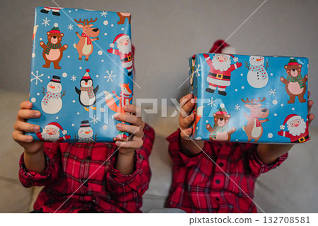 Two cheerful children holding wrapped Christmas presents in front of their faces. Festive holiday concept representing joy, surprise, and gift exchange. 132708581