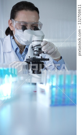 Woman scientist wearing lab coat, white protective gloves and glasses, is using microscope in modern laboratory setup, portrait view. Low key lighting photo. Science and Medicine concept 132708611