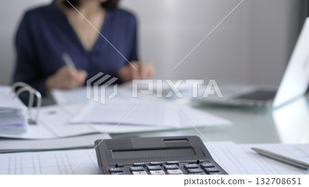 Black calculator, pen and documents are lying on accountant's desk. Businesswoman wearing blue dress is working with documents on the background. Business and audit concept Black calculator, pen and documents are lying on accountant's desk. Businesswoman wearing blue dress is working with documents on the background. Business and audit concept 132708651