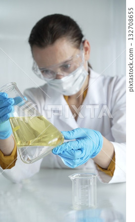Woman scientist, wearing a lab coat, blue protective gloves, is holding an Erlenmeyer flask with yellow oily liquid in laboratory setting, portrait and vertical view. Medicine, science concept Woman scientist, wearing a lab coat, blue protective gloves, is holding an Erlenmeyer flask with yellow oily liquid in laboratory setting, portrait and vertical view. Medicine, science concept 132708655
