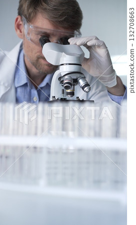 Man scientist wearing lab coat, white gloves and protective glasses using microscope doing research in modern laboratory, portrait view. Low key lighting photo. Science and Medicine Man scientist wearing lab coat, white gloves and protective glasses using microscope doing research in modern laboratory, portrait view. Low key lighting photo. Science and Medicine 132708663