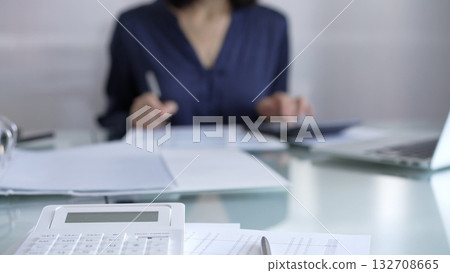 White calculator, pen and documents are lying on accountant's desk. Businesswoman wearing blue dress is working with documents on the background. Business and audit concept White calculator, pen and documents are lying on accountant's desk. Businesswoman wearing blue dress is working with documents on the background. Business and audit concept 132708665