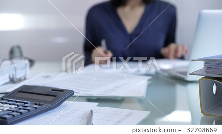 Black calculator, pen and documents are lying on accountant's desk. Businesswoman wearing blue dress is working with documents on the background. Business and audit concept Black calculator, pen and documents are lying on accountant's desk. Businesswoman wearing blue dress is working with documents on the background. Business and audit concept 132708669