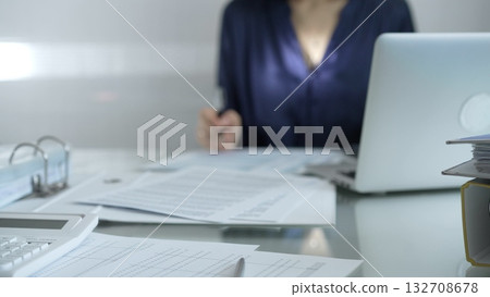Magnifying glass, white calculator, pen and documents are lying on accountant's desk. Businesswoman wearing blue dress is working with documents on the background. Business and audit concept Magnifying glass, white calculator, pen and documents are lying on accountant's desk. Businesswoman wearing blue dress is working with documents on the background. Business and audit concept 132708678