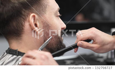 Skilled barber carefully trimming client's beard with precision scissors and sharp comb, working inside contemporary grooming salon Skilled barber carefully trimming client's beard with precision scissors and sharp comb, working inside contemporary grooming salon 132708727
