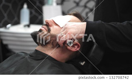 Professional barber grooming client's beard using electric razor, delivering precise trimming in contemporary barbershop setting 132708742