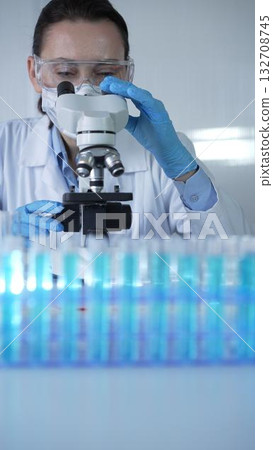 Female scientist wearing lab coat and protective blue gloves using microscope doing research in modern laboratory, portrait view. Low key lighting photo. Science and Medicine 132708745