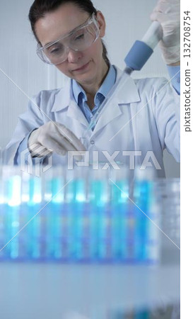 Female scientist wearing lab coat, white medical gloves and protective glasses is using micropipette filling test tubes in modern laboratory. Low key lighting photo. Science concept Female scientist wearing lab coat, white medical gloves and protective glasses is using micropipette filling test tubes in modern laboratory. Low key lighting photo. Science concept 132708754