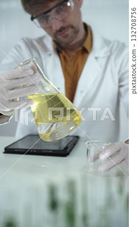 Man scientist wearing white protective gloves, and glasses is holding a yellow chemical solution inside an Erlenmeyer flask in a laboratory, vertical portrait. Science and medicine 132708756
