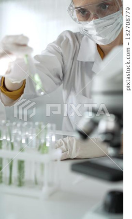 Woman scientist wearing lab coat, white gloves, face mask and protective glasses, is holding a test tube with plants inside near microscope in laboratory, vertical portrait view. Science and medicine 132708779