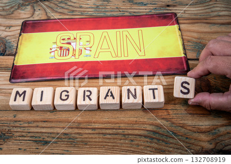 Spanish flag with wooden blocks spelling MIGRANTS. Symbolizing migration, society and national Spanish flag with wooden blocks spelling MIGRANTS. Symbolizing migration, society and national 132708919
