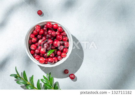 Fresh cranberries in a white bowl on a light background with green branch and sunlight. Fresh cranberries in a white bowl on a light background with green branch and sunlight. 132711182
