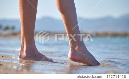 Close up of female feet walking barefoot on white grainy sand of golden beach on blue ocean water background Close up of female feet walking barefoot on white grainy sand of golden beach on blue ocean water background 132711418