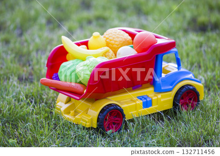 Bright plastic colorful toy car truck carrying basket with toy fruits and vegetables outdoors on sunny summer day. 132711456