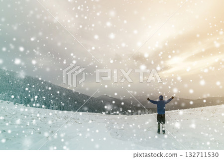 Back view of tourist hiker in warm clothing with backpack standing with raised arms on clearing covered with snow on spruce forest mountain and cloudy sky copy space background. Back view of tourist hiker in warm clothing with backpack standing with raised arms on clearing covered with snow on spruce forest mountain and cloudy sky copy space background. 132711530