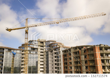 Apartment or office tall building under construction. Brick walls, glass windows, scaffolding and concrete support pillars. Tower crane on bright blue sky copy space background. Apartment or office tall building under construction. Brick walls, glass windows, scaffolding and concrete support pillars. Tower crane on bright blue sky copy space background. 132711574