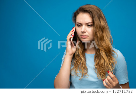 Young woman with serious facial expression talking on mobile phone against blue background Young woman with serious facial expression talking on mobile phone against blue background 132711782