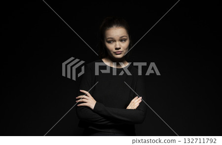 Young woman with skeptical expression and crossed arms posing in dark studio Young woman with skeptical expression and crossed arms posing in dark studio 132711792