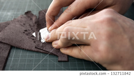 Artisan preparing brown leather edges for gluing in handmade leather crafting process 132711842