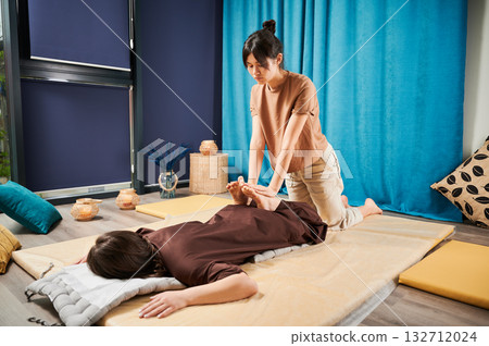 Masseuse making thai yoga massage. Woman therapist doing traditional massage treatment, stretching female patient's body. Client lying on mat, enjoying hand massage in calm atmosphere. 132712024