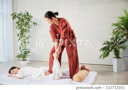 Masseuse making thai yoga massage. Woman therapist doing traditional massage treatment for female patient indoors. Client lying on mat, meditating, enjoying hand massage in calm atmosphere. 132712031
