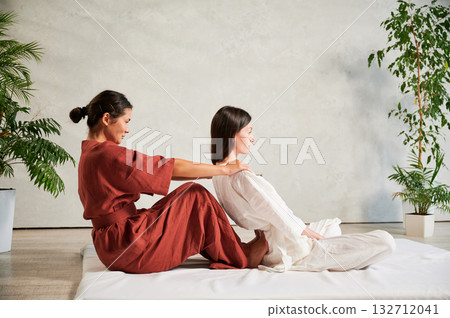 Masseuse making thai yoga massage. Woman therapist doing traditional massage treatment, stretching female patient's body. Client sitting on mat, enjoying hand massage in calm atmosphere. 132712041