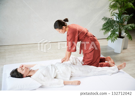 Masseuse making thai yoga massage. Woman therapist doing traditional massage treatment, stretching female patient's body. Client lying on mat, enjoying hand massage in calm atmosphere. 132712042