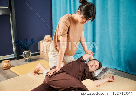 Masseuse making thai yoga massage. Woman therapist doing traditional massage treatment, stretching female patient's body. Client lying on mat, enjoying hand massage in calm atmosphere. 132712044