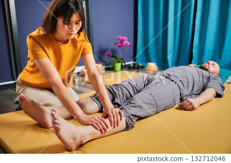 Masseuse making thai yoga massage. Woman therapist doing traditional massage treatment for male patient indoors. Bald client lying on mat, meditating, enjoying hand massage in calm atmosphere. 132712046