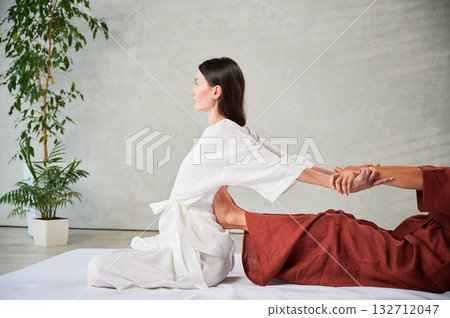 Masseuse making thai yoga massage. Woman therapist doing traditional massage treatment, stretching female patient's body. Client sitting on mat, enjoying hand massage in calm atmosphere. 132712047