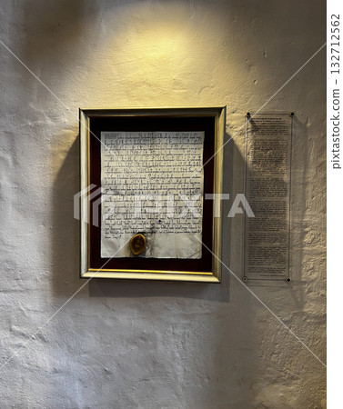 An illuminated framed medieval document with seal hangs on a textured wall in Osnabruk, supported by a modern explanation panel. An illuminated framed medieval document with seal hangs on a textured wall in Osnabruk, supported by a modern explanation panel. 132712562