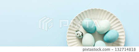 A collection of decorated blue Easter eggs sits on a white plate, showcasing various shades and patterns. The light background enhances the festive atmosphere of Easter, banner 132714818