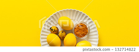 Bright yellow Easter eggs arranged on a white plate with quail eggs and a small bowl of turmeric add a vibrant touch to the holiday decorations and festivities, a way to dye eggs yellow, banner 132714822