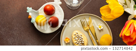 A beautifully arranged Easter table setting showcases a yellow napkin wrapped around a red apple, surrounded by vibrant flowers and a festive bunny decoration, banner 132714842