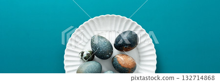 Decorative Easter eggs with various textures and colors are arranged neatly on a white plate, resting on a vibrant teal surface, creating a festive spring atmosphere, banner 132714868