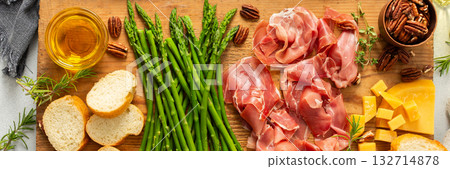 A beautifully arranged charcuterie board features fresh asparagus, various meats, cheeses, and nuts, set on a wooden plank with slices of bread. Perfect for sharing at gatherings, banner 132714878