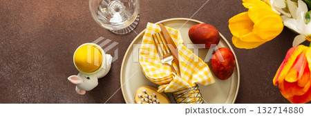 A beautifully arranged Easter table setting showcases a yellow napkin wrapped around a red apple, surrounded by vibrant flowers and a festive bunny decoration, banner 132714880