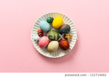 A collection of vibrant Easter eggs in various colors is artfully arranged on a white plate, set against a soft pink background, capturing the essence of the holiday's decorations 132714882